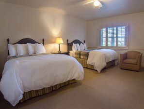 A hotel room with two beds, a nightstand with a lamp, a chair, and a window with blinds.