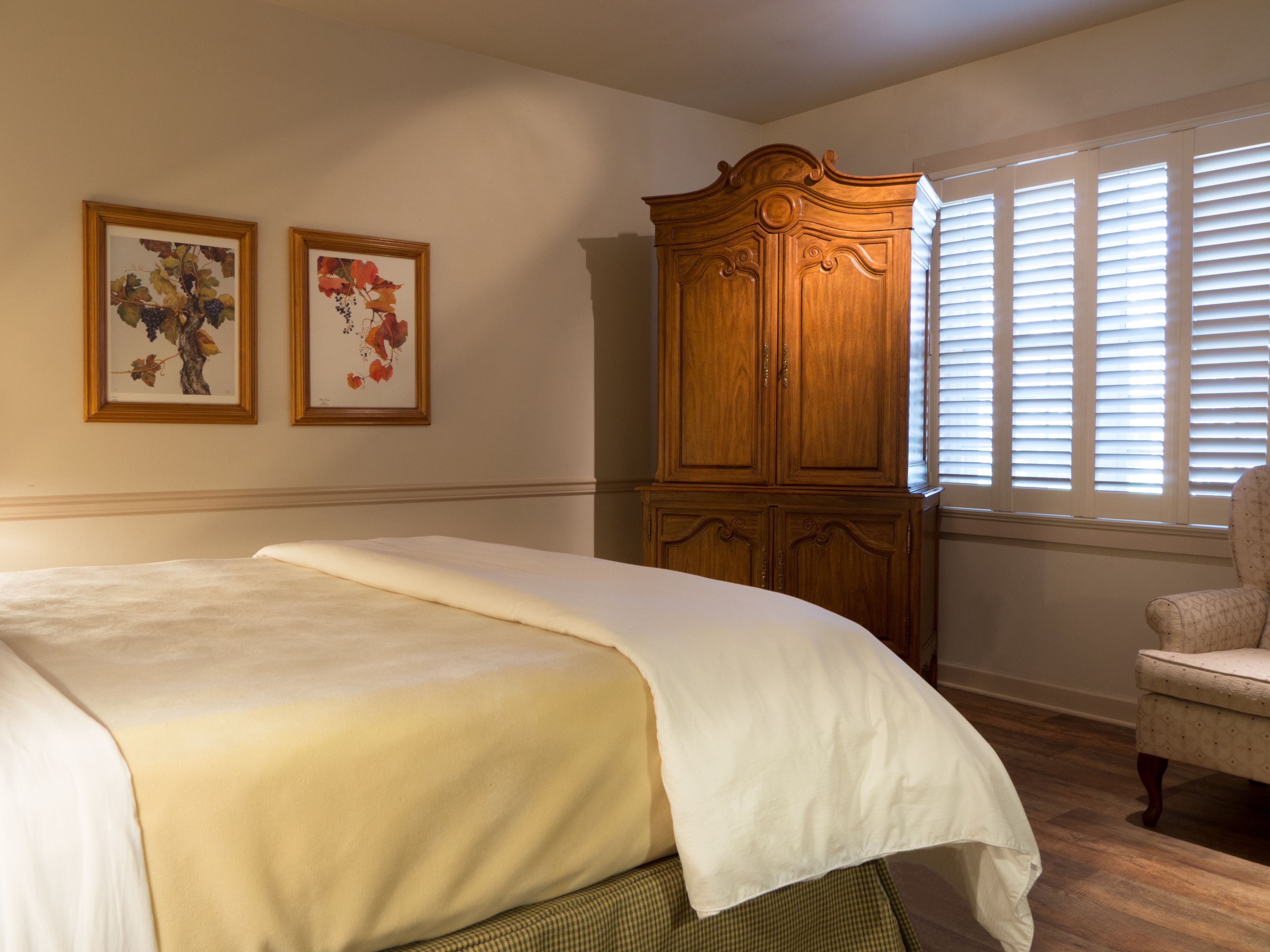 A bedroom with a large bed, a wooden wardrobe, two framed floral paintings, and a window with blinds.