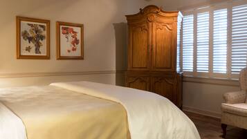 A bedroom with a large bed, a wooden wardrobe, two framed floral paintings, and a window with blinds.