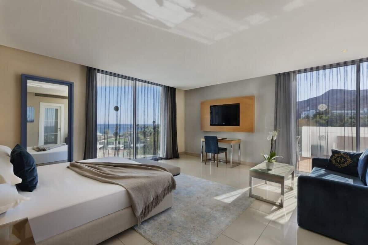A modern hotel room with a large bed, a desk, a sofa, a TV, and a balcony with a view.