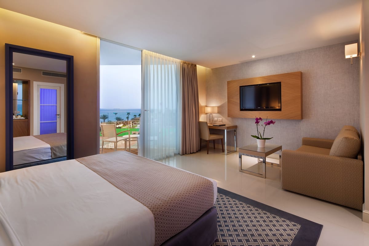 A hotel room with a bed, a desk, a sofa, a TV mounted on the wall, and a view of the ocean.