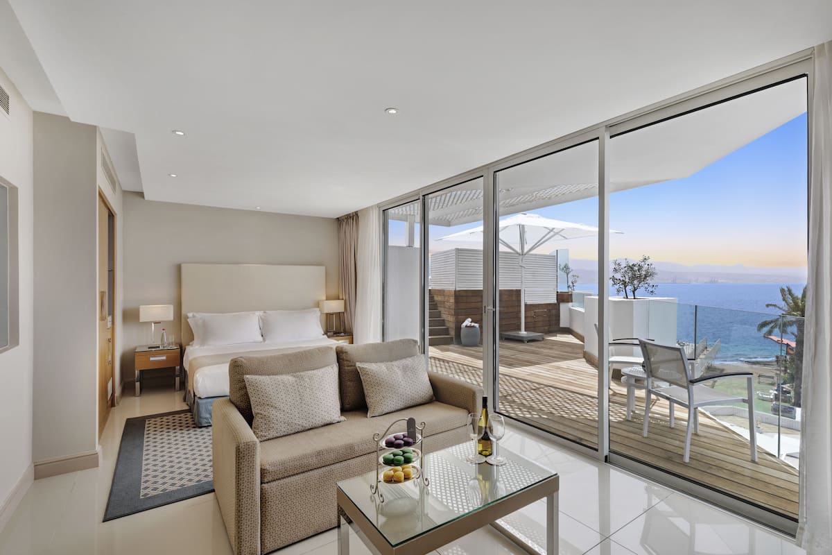 A modern hotel room with a large bed, a sofa, a glass coffee table, and a balcony with a view of the sea.