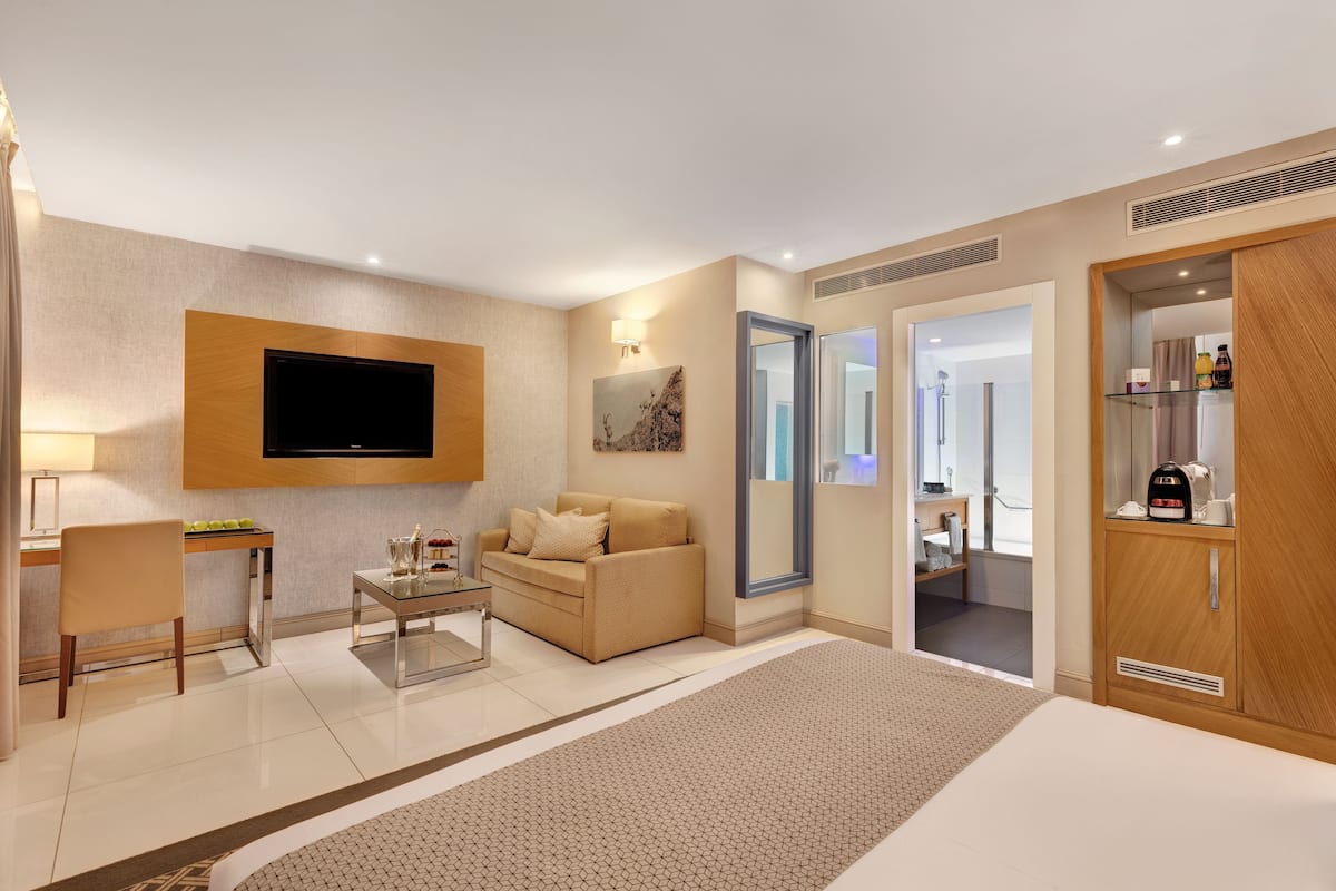 A modern hotel room with a flat-screen TV, a sofa, a coffee table, a desk, and a wardrobe.