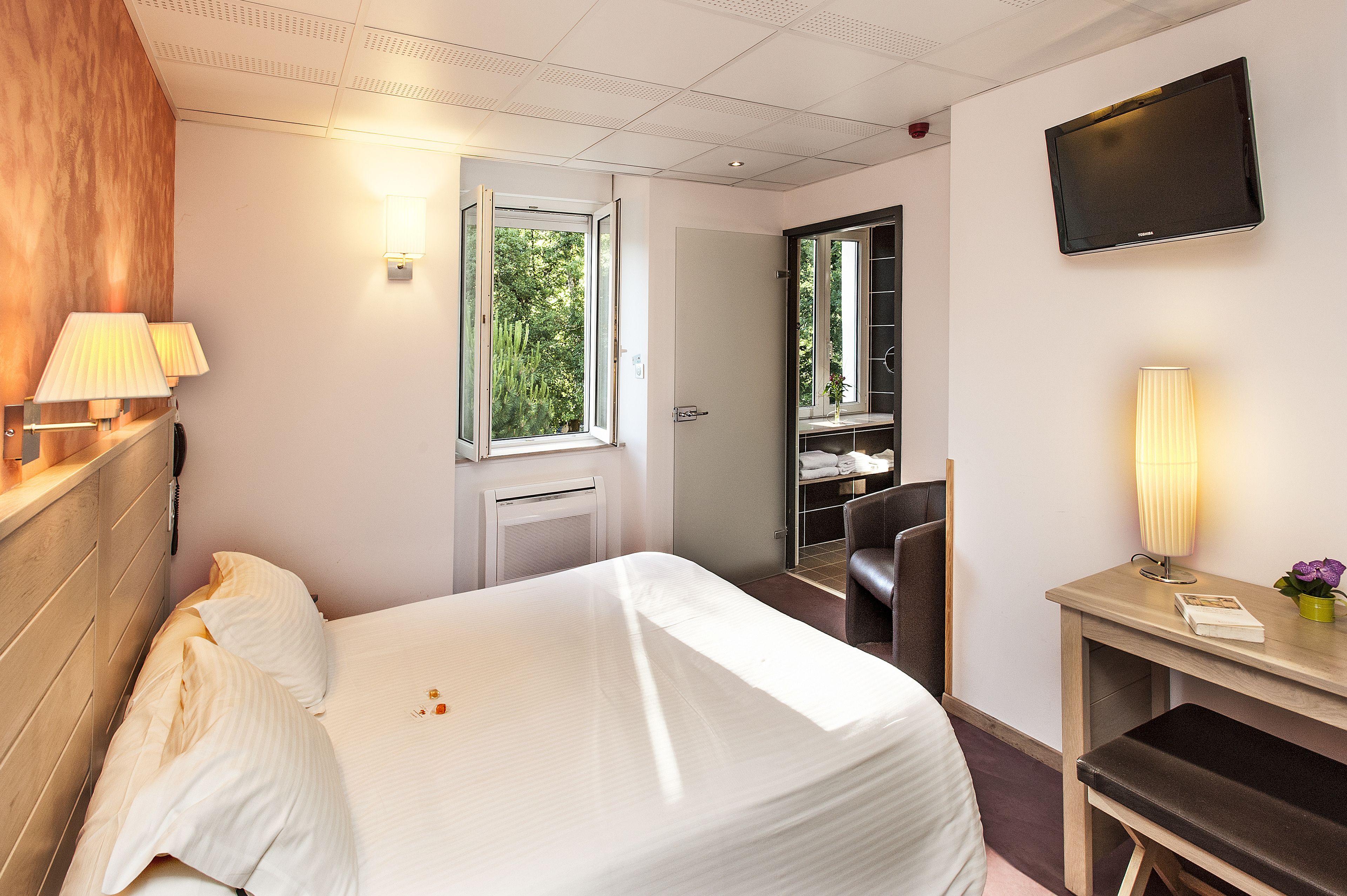 Habitación de hotel con cama, lámparas de noche, un escritorio con silla, televisor y ventana con vista a follaje.