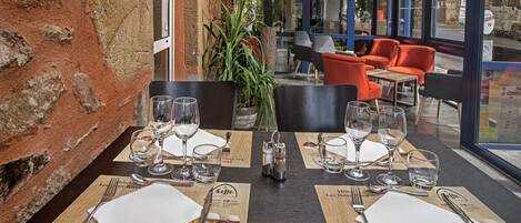 A restaurant with tables set for two, featuring a rustic brick wall and large windows offering a view of the street.