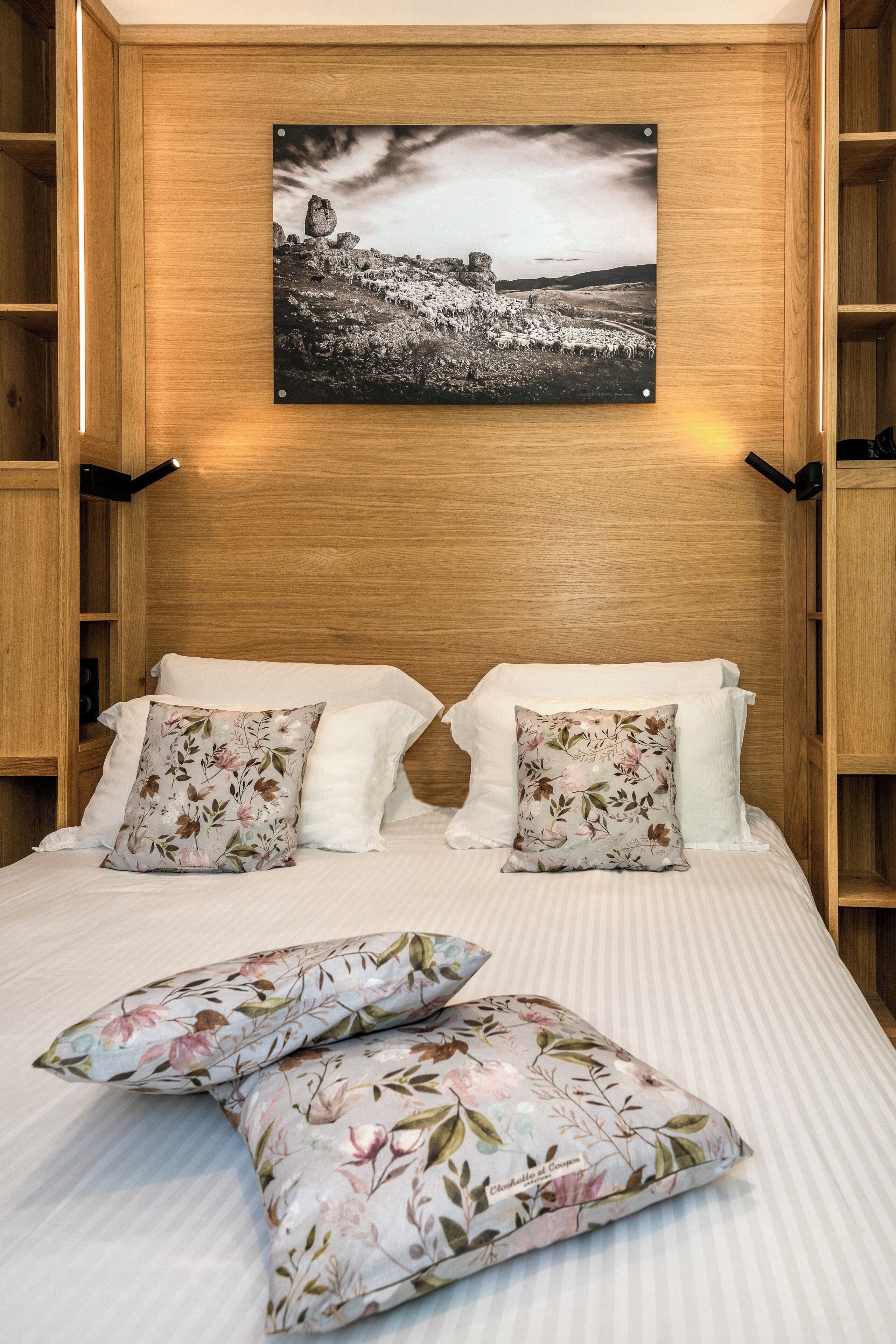A bedroom with a wooden headboard, a framed landscape photo, and floral-patterned pillows.