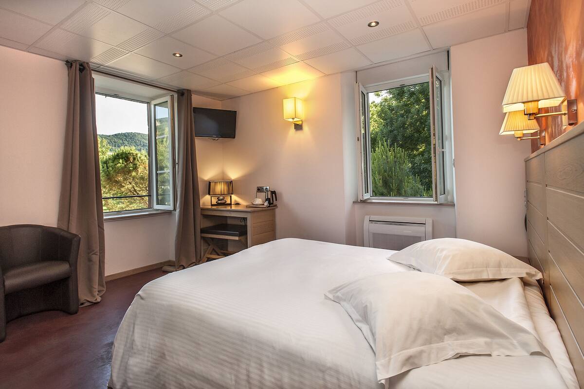 A hotel room with a bed, a desk with a lamp, a chair, a TV, and windows with curtains.