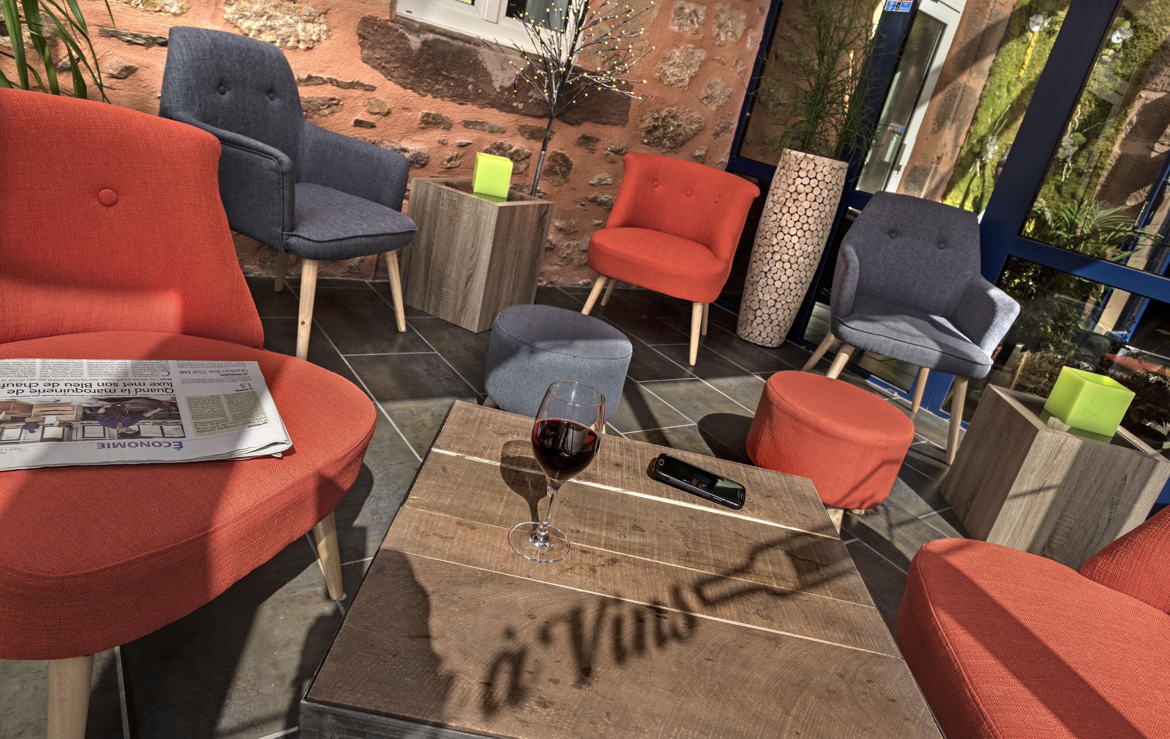 A cozy outdoor seating area with red and grey upholstered chairs, a wooden coffee table with a glass of red wine, and a newspaper.