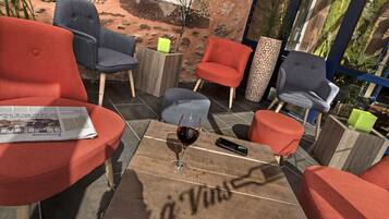 A cozy outdoor seating area with red and grey upholstered chairs, a wooden coffee table with a glass of red wine, and a newspaper.