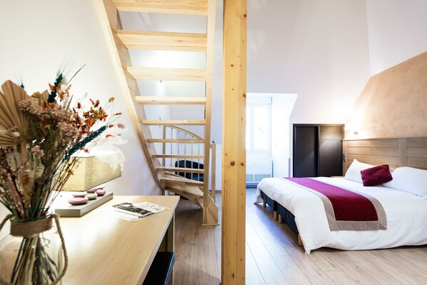 Una habitación con escalera de madera, cama con una manta roja y escritorio con un florero de flores secas.