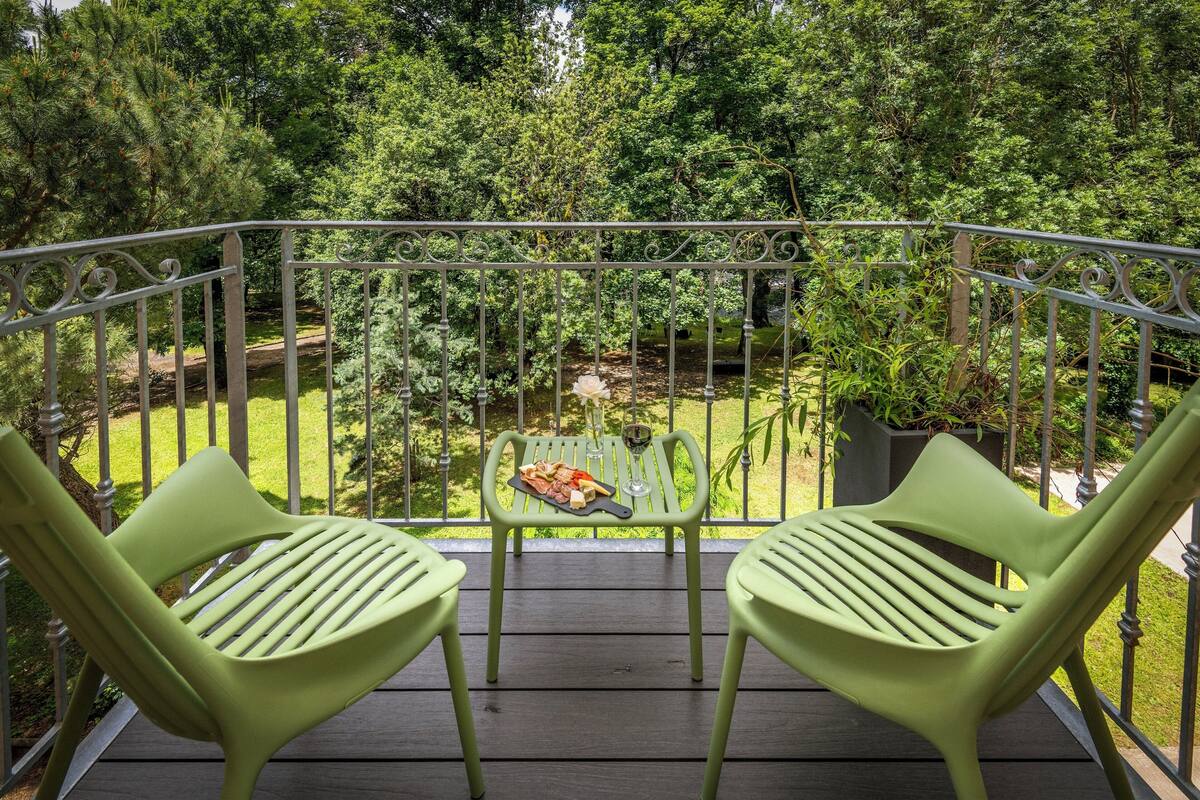 A balcony with two green chairs, a small table with food and a glass, and a view of a garden with trees and a pond.