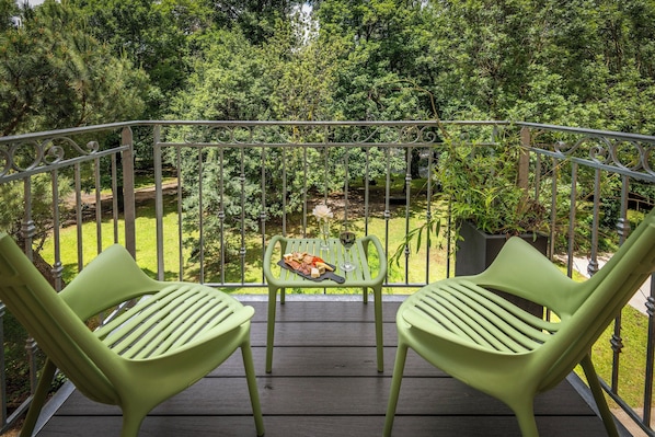A balcony with two green chairs, a small table with food and a glass, and a view of a garden with trees and a pond.