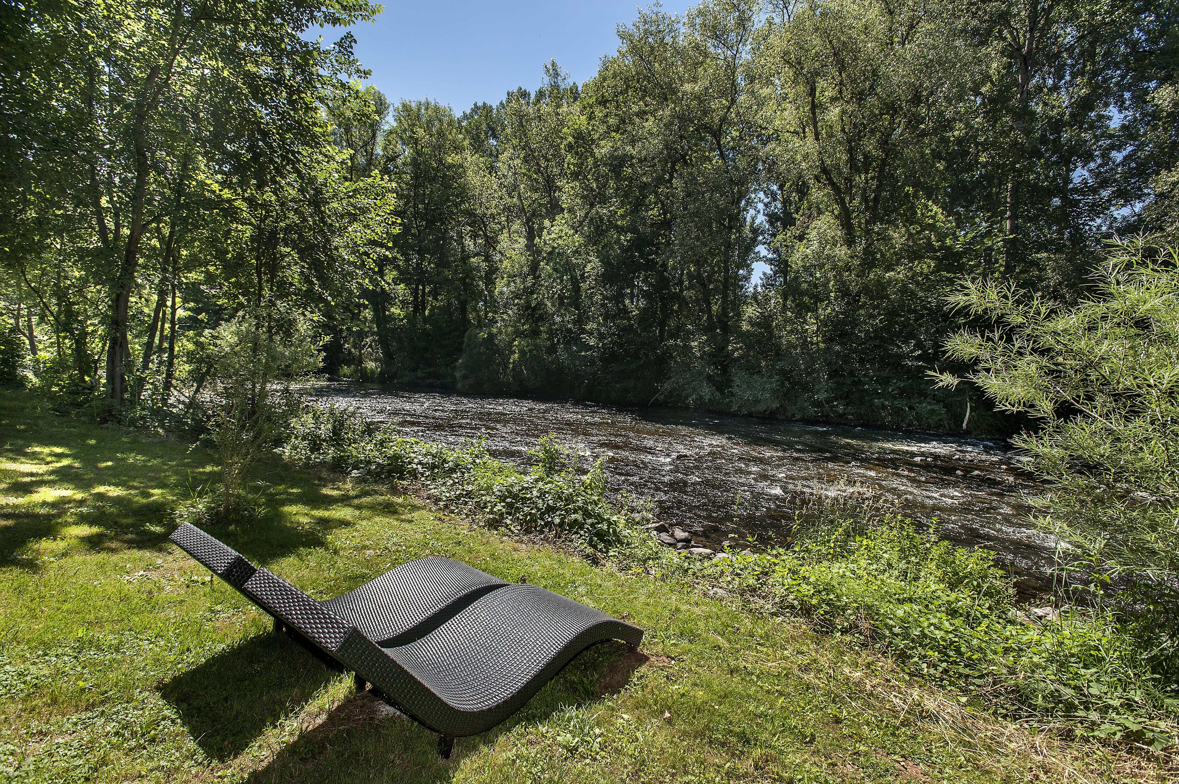 Una zona junto al río con una silla de descanso y un bosque denso al fondo.