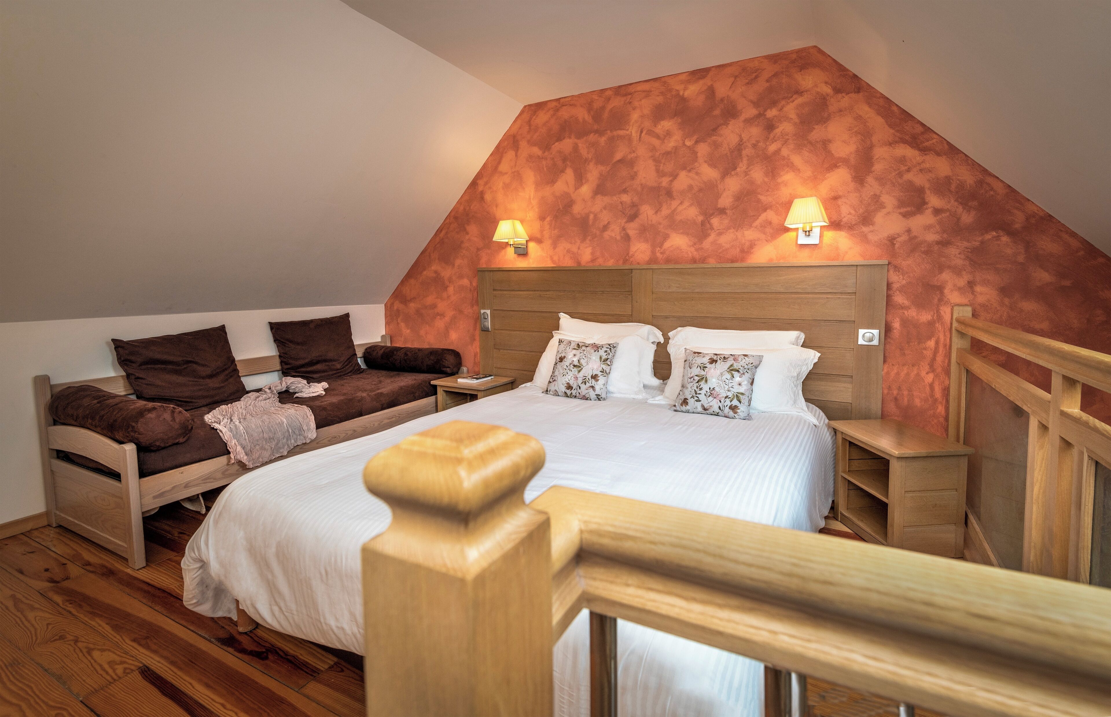 Una camera da letto con soffitto spiovente, un letto con biancheria bianca, due abat-jour, un comodino in legno e una panca di legno con cuscini.