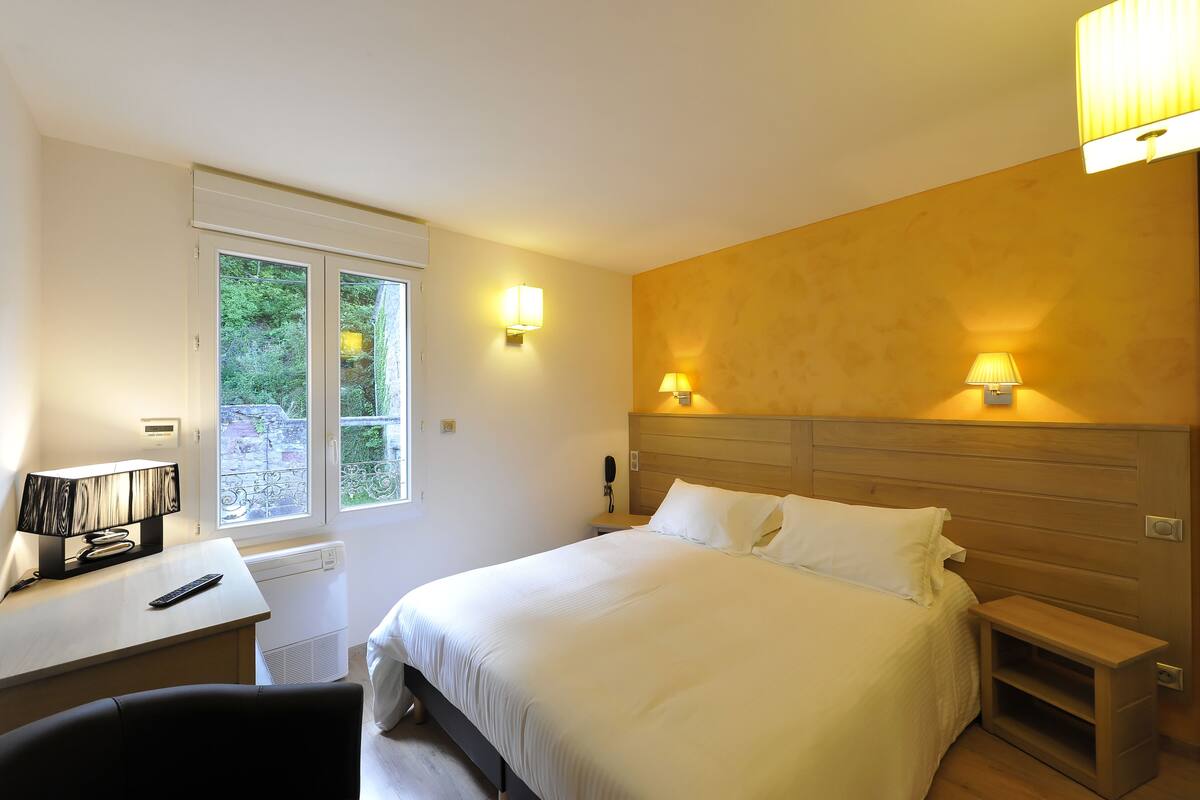 A bedroom with a bed, bedside lamps, a desk with a lamp, and a window with a view of greenery.