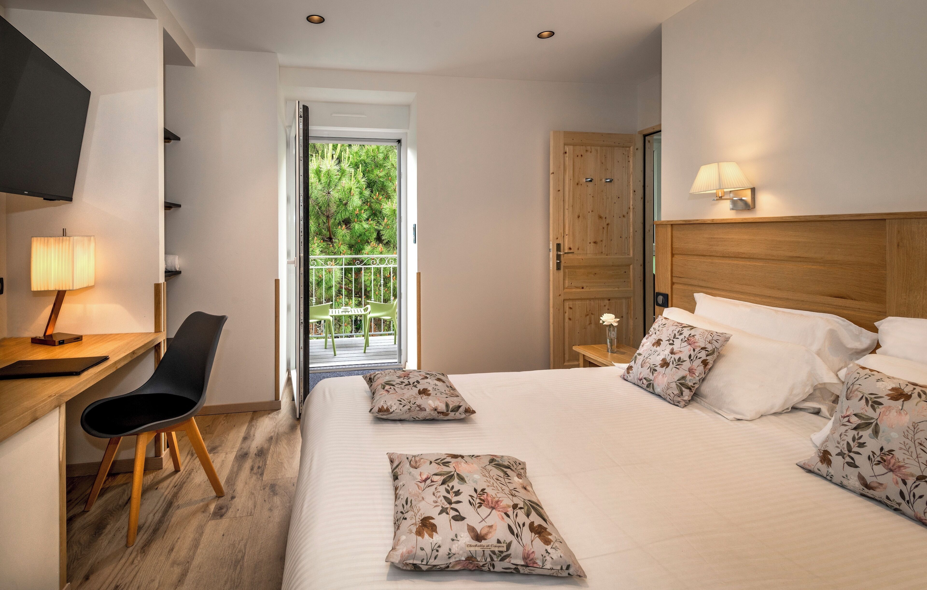 Hotelkamer met een bed, bureau met stoel, televisie, houten garderobe en een balkon met beplanting.