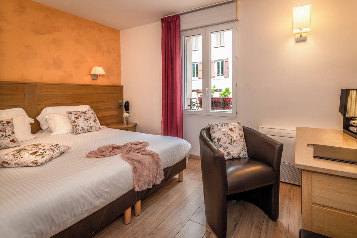 A hotel room with a bed, a desk, a chair, a window with curtains, and a lamp.