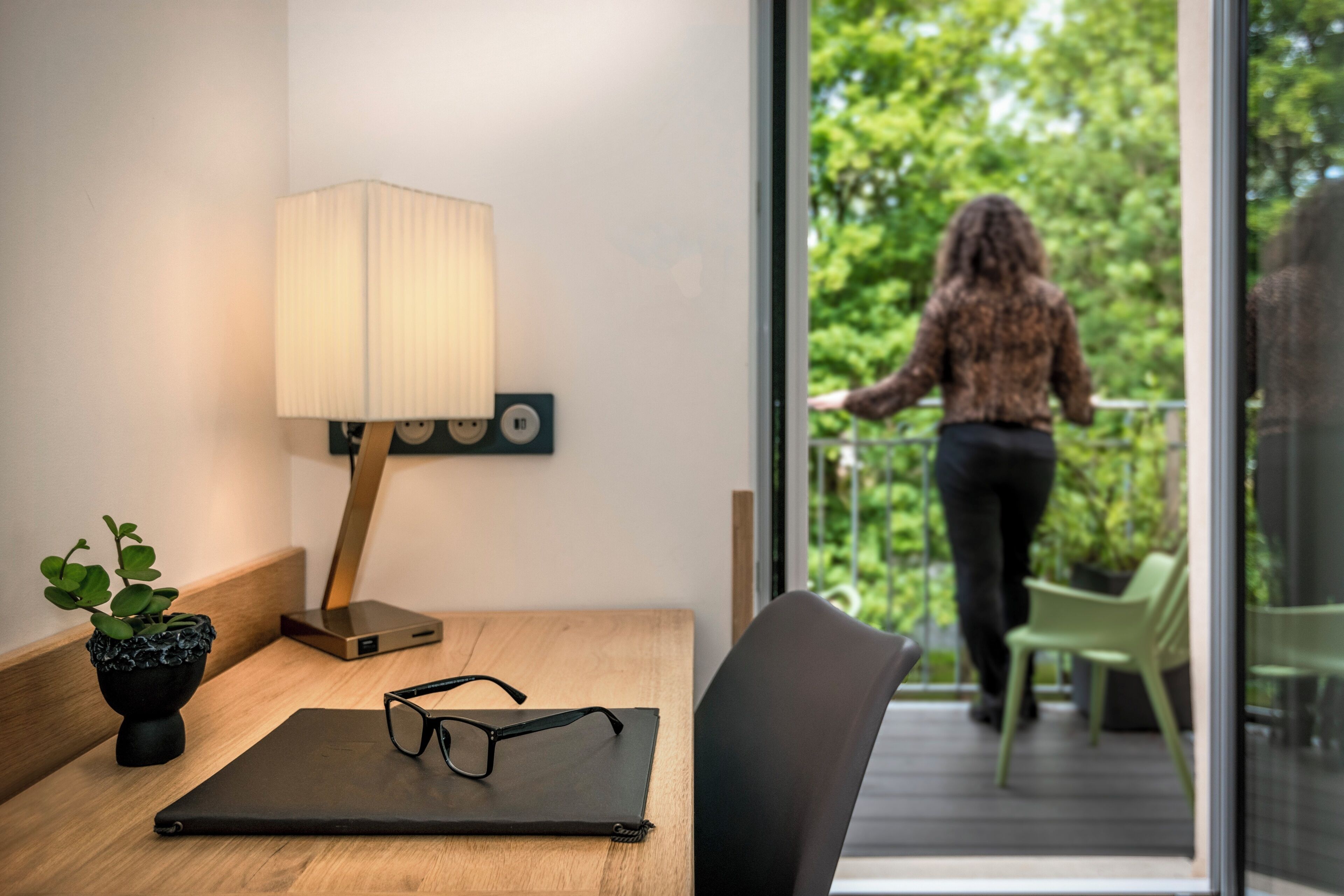 Una habitación moderna con un escritorio de madera, una lámpara, un par de gafas y vistas a un balcón con plantas.