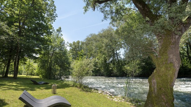 Zona de relaxació al costat del riu, amb una cadira de relax i una taula petita, envoltada de vegetació abundant i un arbre gran.