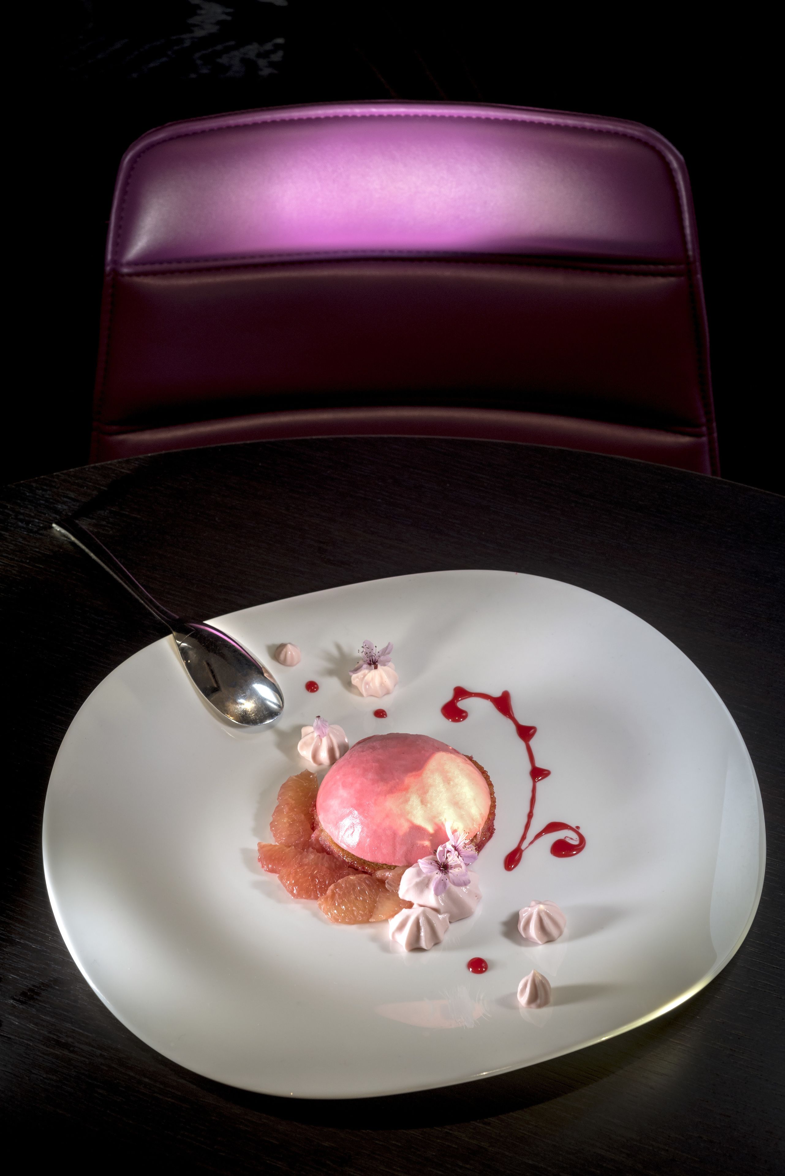 Un dessert servi dans une assiette blanche, accompagné d’une cuillère, est posé sur une table dont le plateau est en bois foncé.