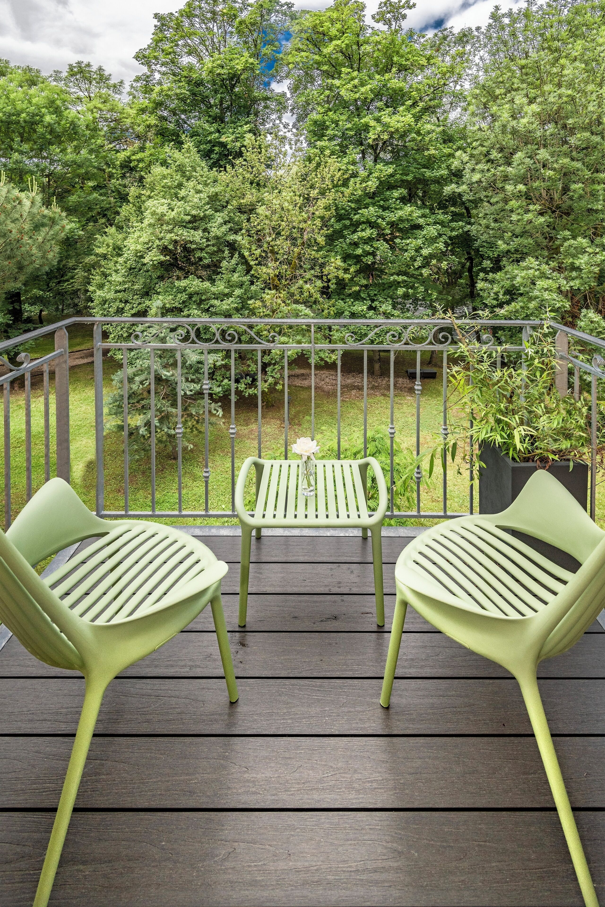 Une terrasse en bois avec deux chaises vertes et une petite table, entourée d’une rambarde en métal, donnant sur un jardin luxuriant.