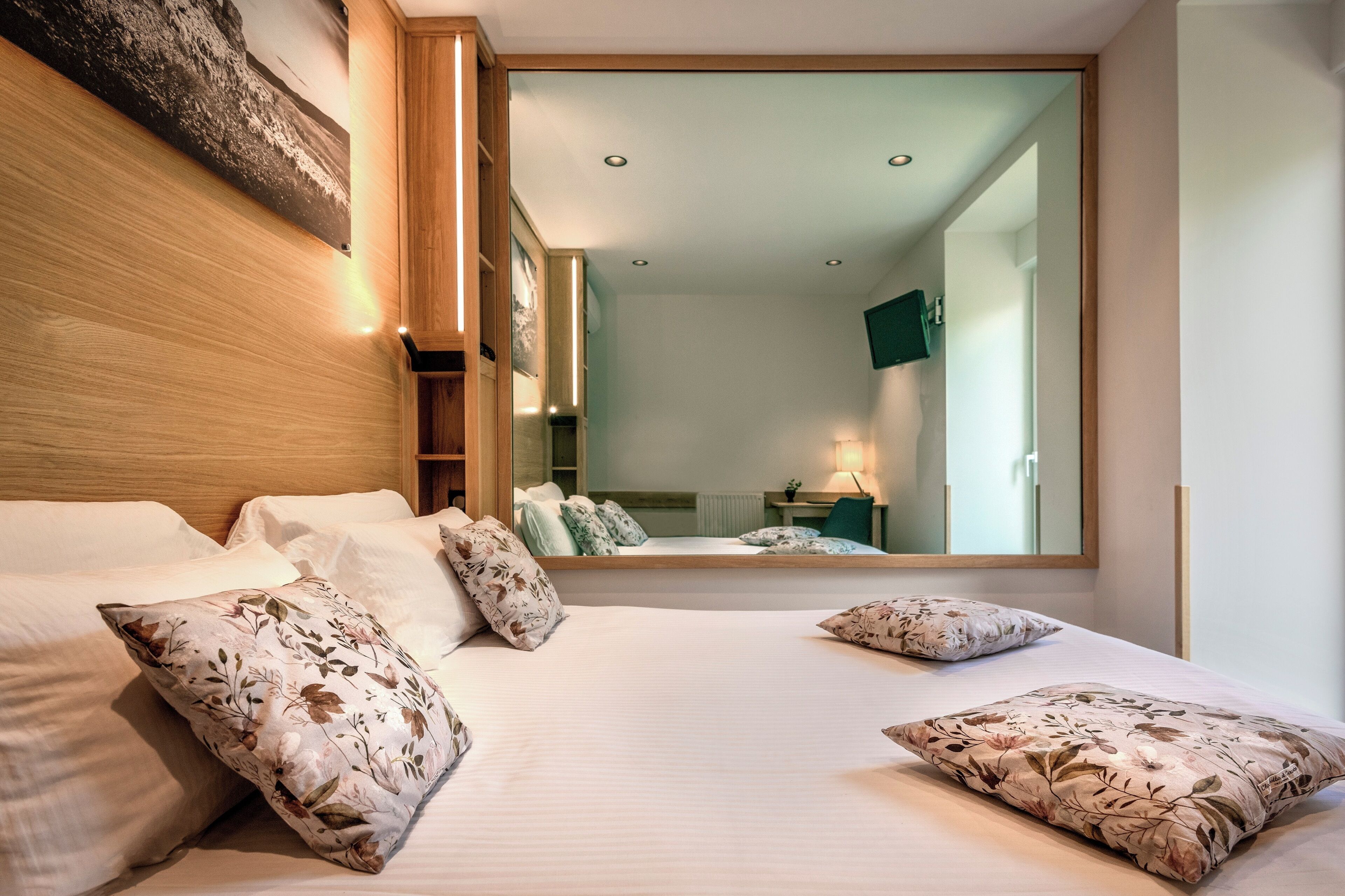 Una habitación de hotel moderna con una cama grande, almohadas estampadas con motivos florales, un cabecero de madera, un espejo amplio y vistas de la habitación contigua.