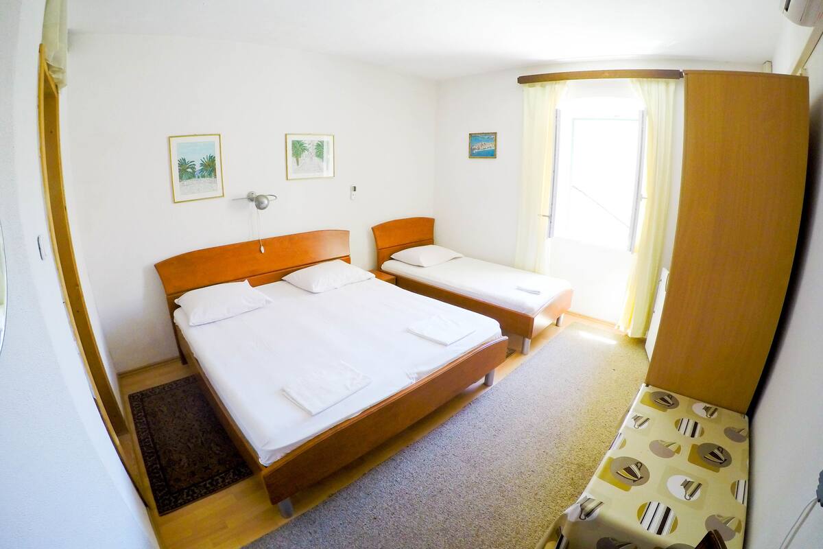 Quarto de hotel com duas camas de solteiro, cabeceira de madeira, uma secretária pequena, uma cadeira e uma janela com cortinas.