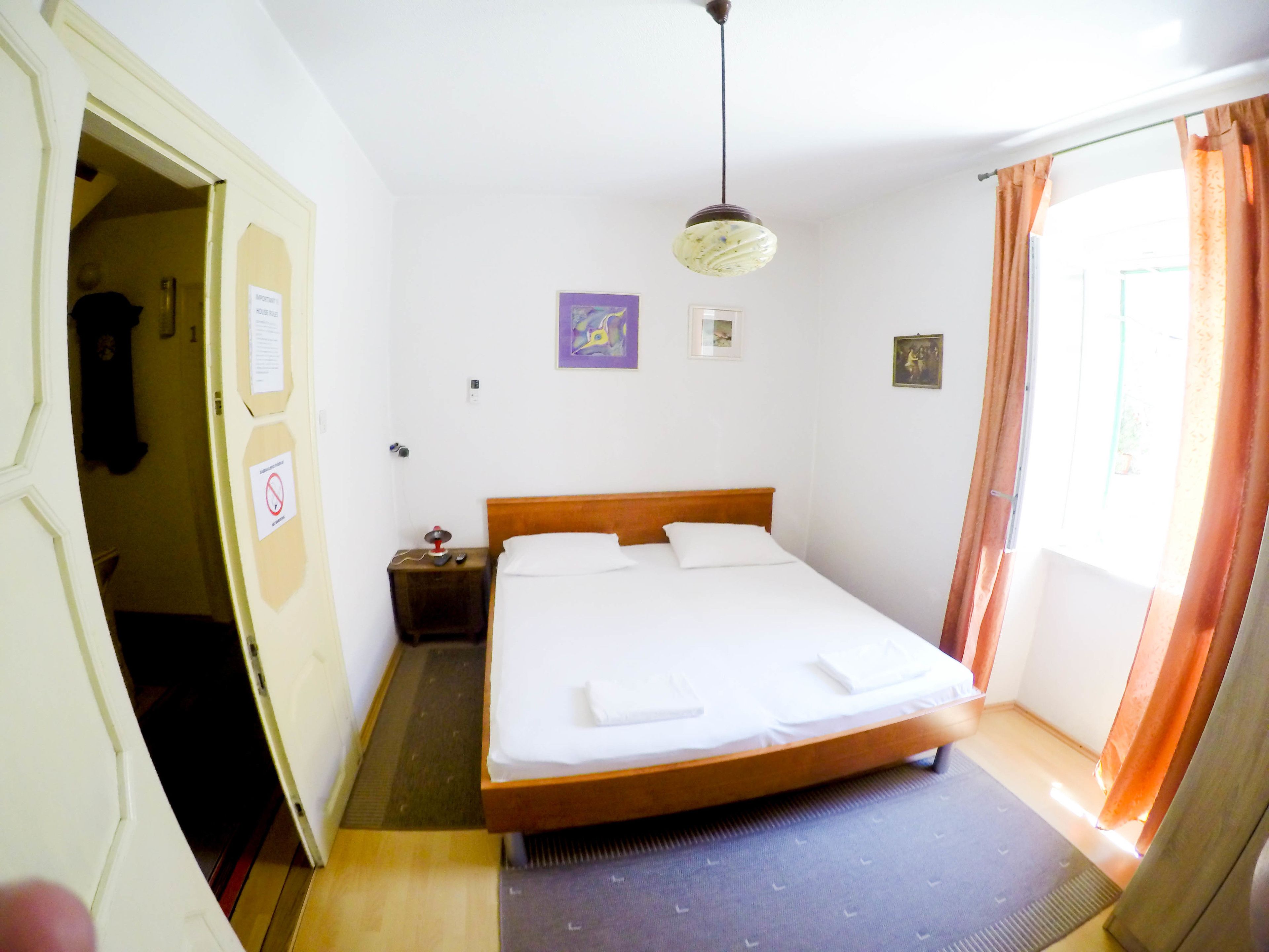 A bedroom with a single bed, a bedside table, a hanging light fixture, and a door leading to another room.