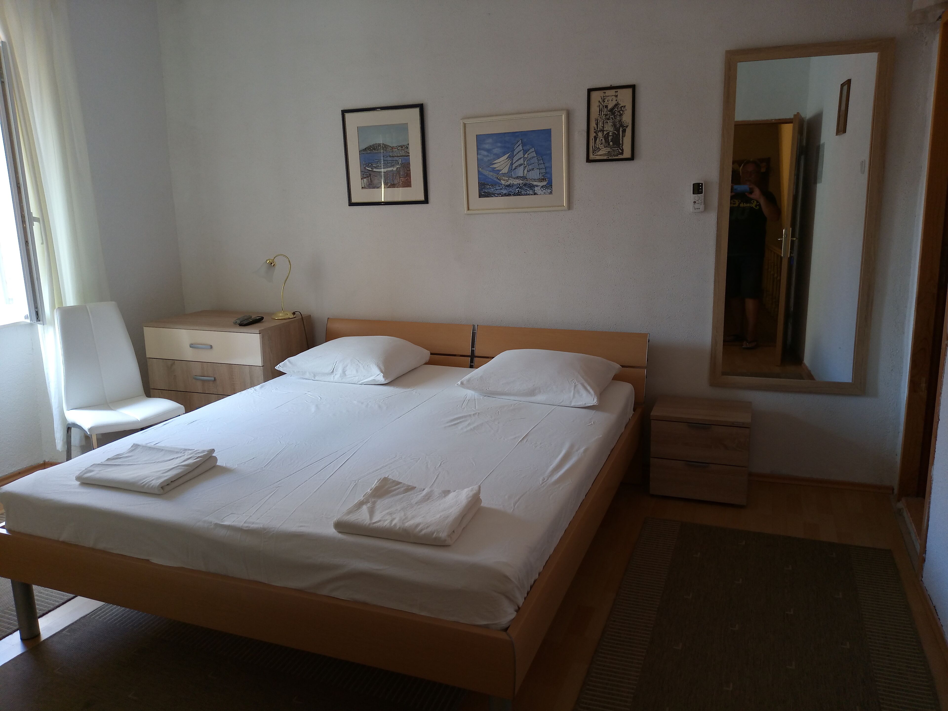 Una cama bien hecha con sábanas y almohadas blancas, una mesita de noche con lámpara, una silla y un aparador. Hay tres cuadros en la pared.