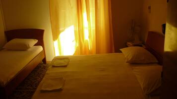 A hotel room with two single beds, a bedside table, a chair, and a standing fan.