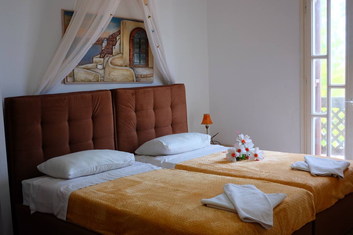 Una cama doble con cabecera marrón, almohadas blancas y colcha amarilla. Encima de la cama hay un cuadro y una ventana con vistas a zonas verdes.