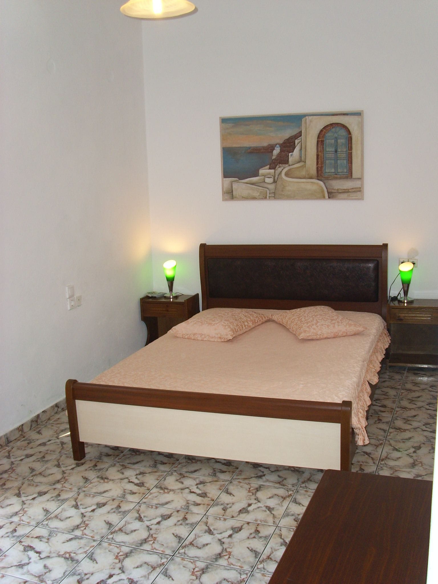 A bedroom with a bed, two bedside tables, a nightstand, a painting on the wall, and a tiled floor.