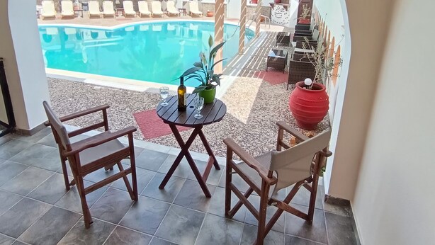 A view through an arched doorway of a pool area with a tiled floor, two wooden chairs, a small table with a bottle and glasses, and a red pot. - Sweet Heart Studio - Apartments (Santorini)