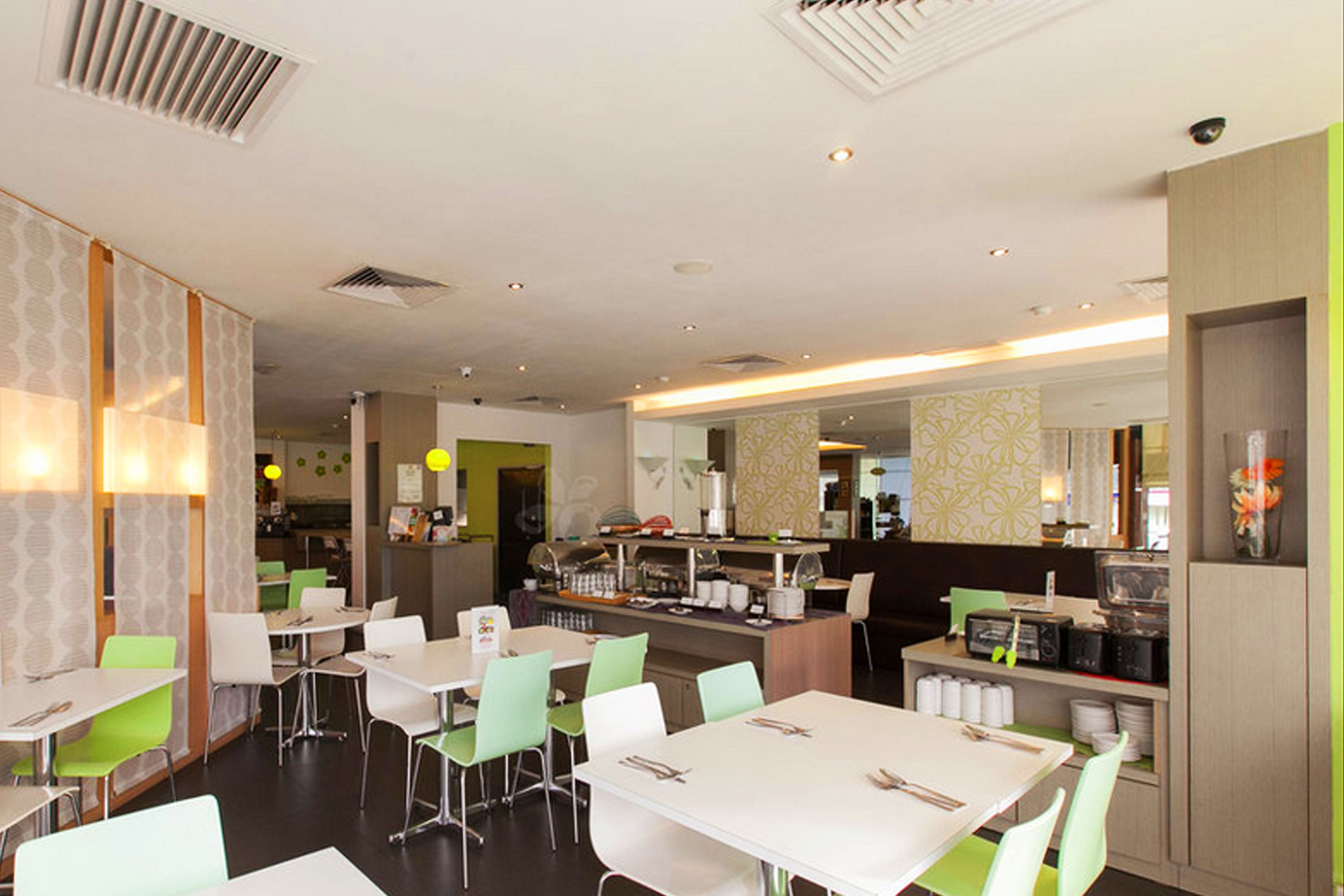 A modern restaurant with white tables and green chairs, a counter with a cash register, and a patterned wall.