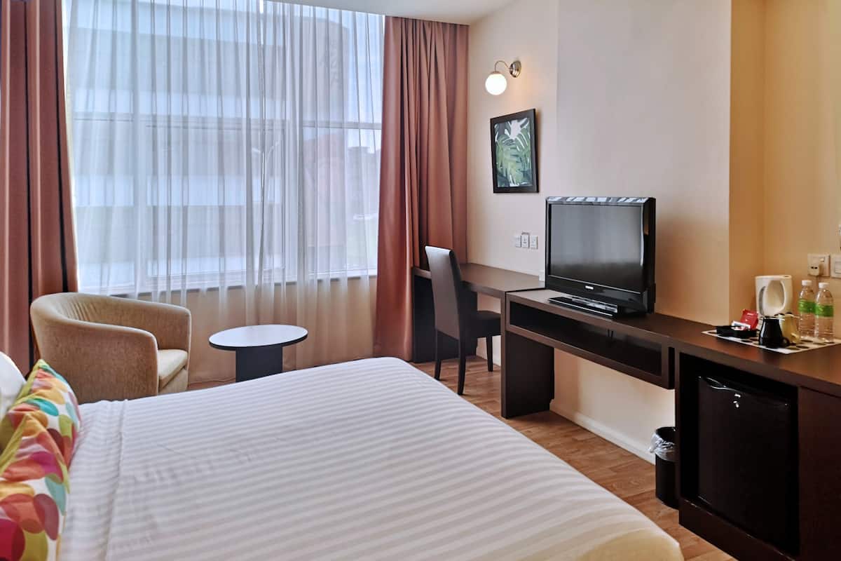 Una habitación de hotel con cama, escritorio con televisor, silla, mesita y ventana con cortinas.