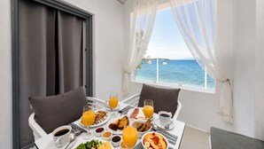 A breakfast table with a view of the sea, a window with sheer curtains, and a balcony with a white railing. - Blue Waves Suites & Apartments To Kyma (Paros)