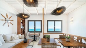A modern living room with a sofa, dining table, chairs, and a view of the sea through large windows. - Blue Waves Suites & Apartments To Kyma (Paros)