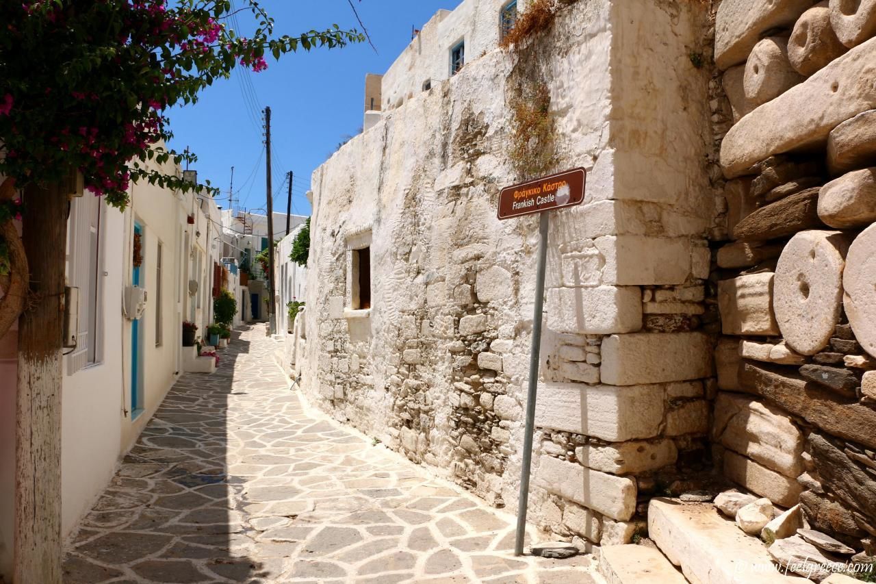 A narrow cobblestone street with stone buildings, a sign for Frankish Castle, and a tree with pink flowers.