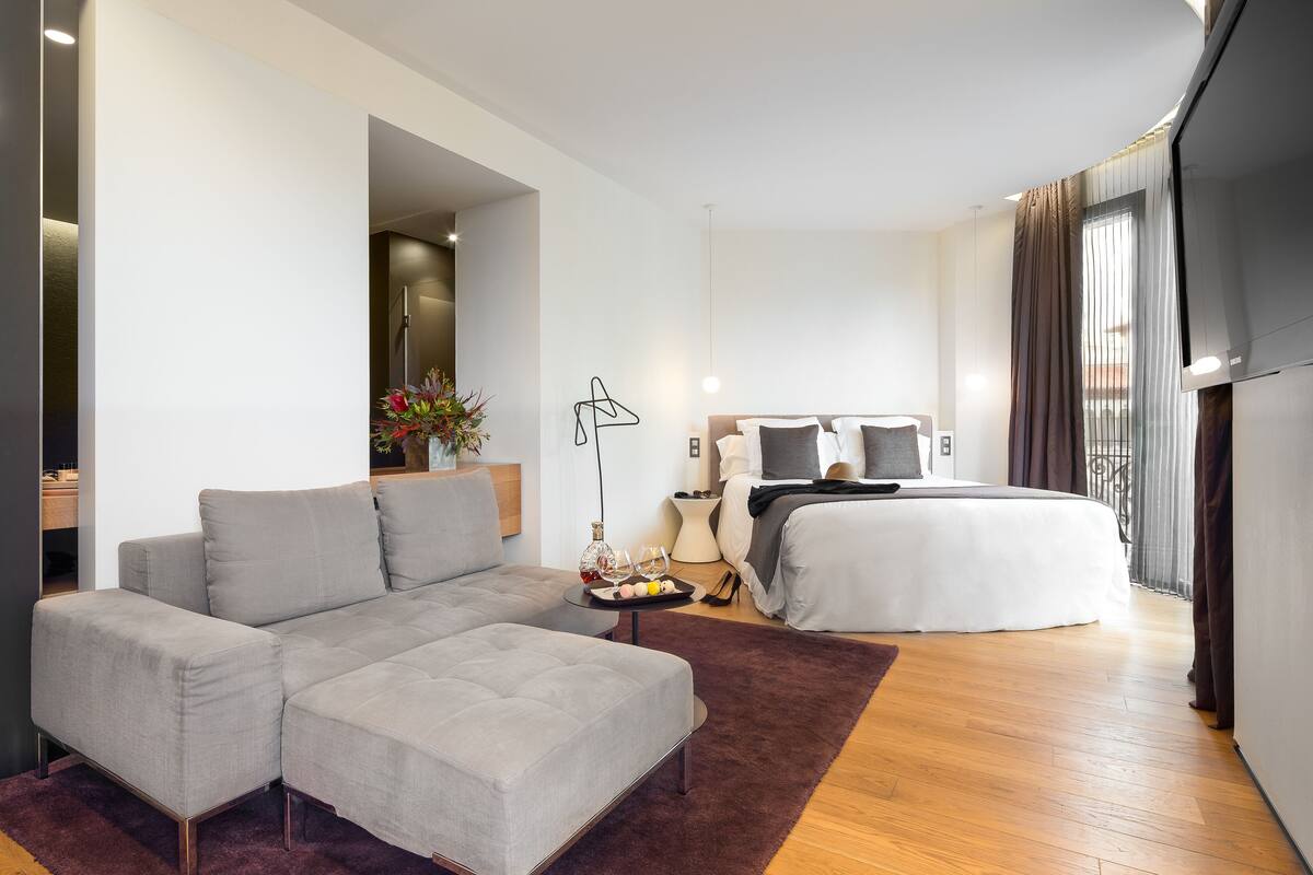 A modern hotel room with a grey sofa, a wooden floor, a large bed, and a TV.