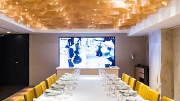 A long dining table set with white tableware and yellow chairs, with a large screen displaying a chef in the background.