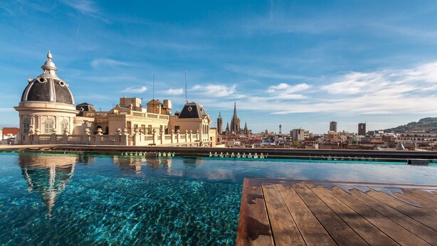 A clear pool with a wooden deck in the foreground, a historic building with a dome and classical architecture in the background, and a cityscape with various buildings and a church.