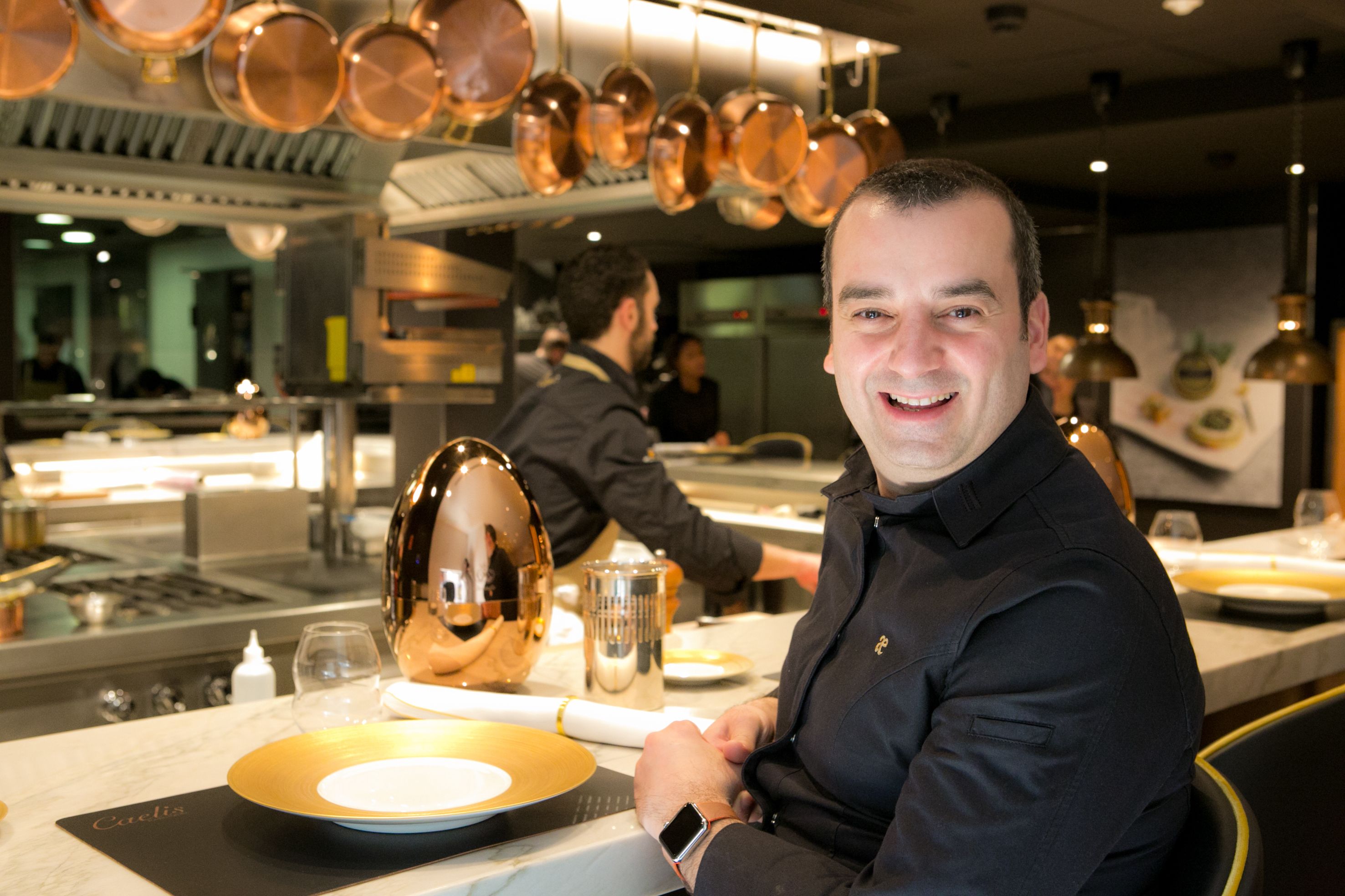 A chef in a professional kitchen with copper cookware, stainless steel appliances, and a marble countertop.