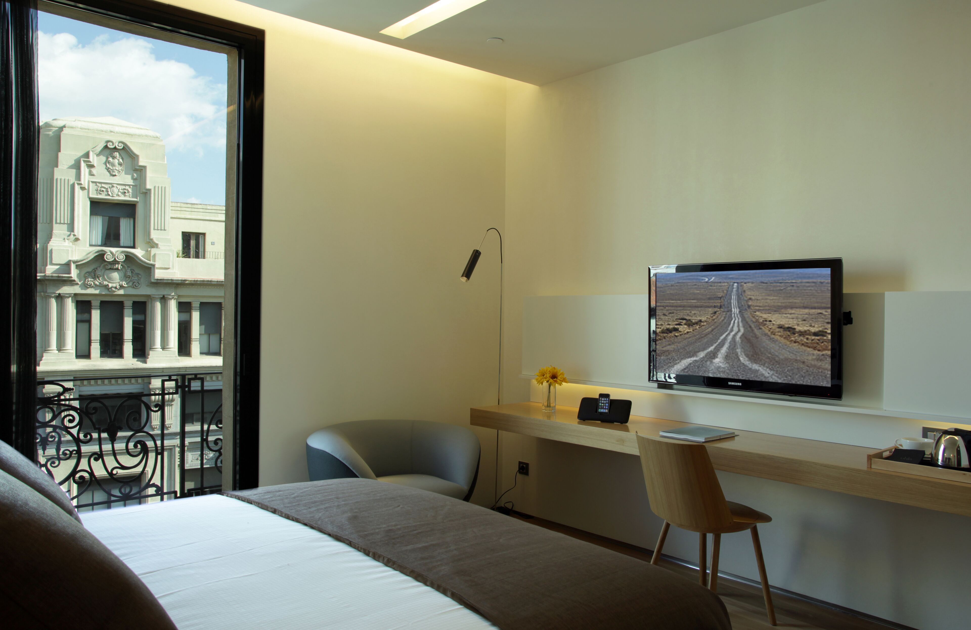 Habitación de hotel con cama, escritorio con computadora, silla, televisor en la pared y ventana con vista a un edificio.