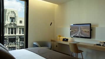 Habitación de hotel con cama, escritorio con computadora, silla, televisor en la pared y ventana con vista a un edificio.