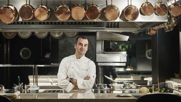 A chef in a professional kitchen with copper cookware hanging above and various kitchen utensils and ingredients on the counter.