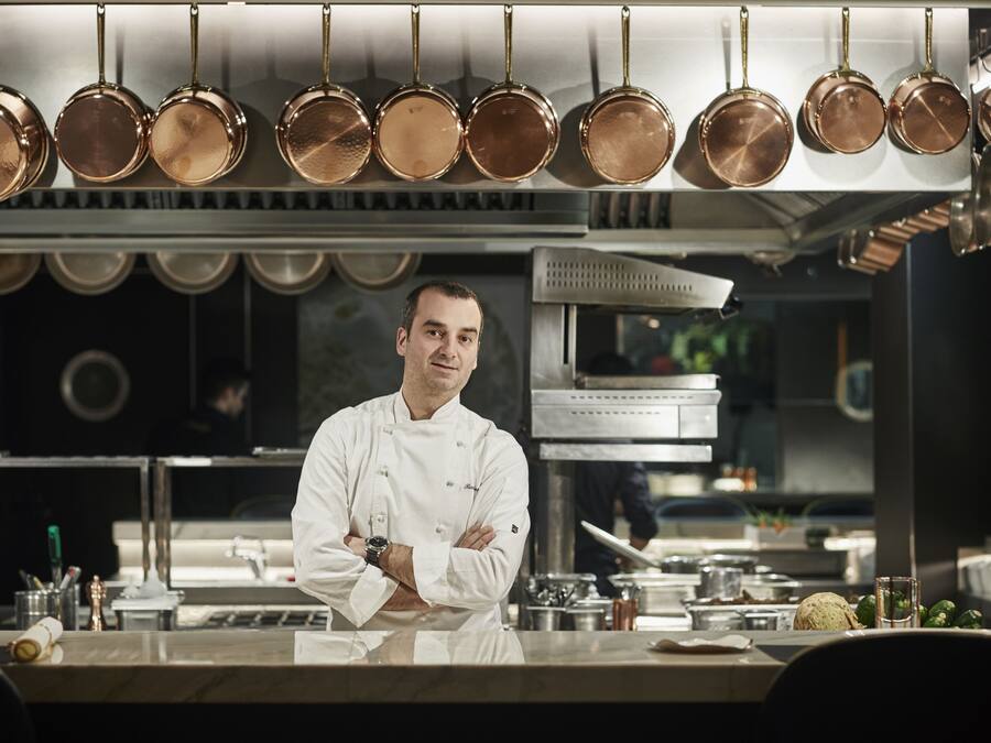 Een kok in een professionele keuken, met koperen pannen erboven en diverse keukengerei en ingrediënten op het aanrecht.