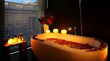 Une salle de bain avec une baignoire remplie de pétales de rose, un vase avec des fleurs rouges et des bougies posées sur une table d’appoint.