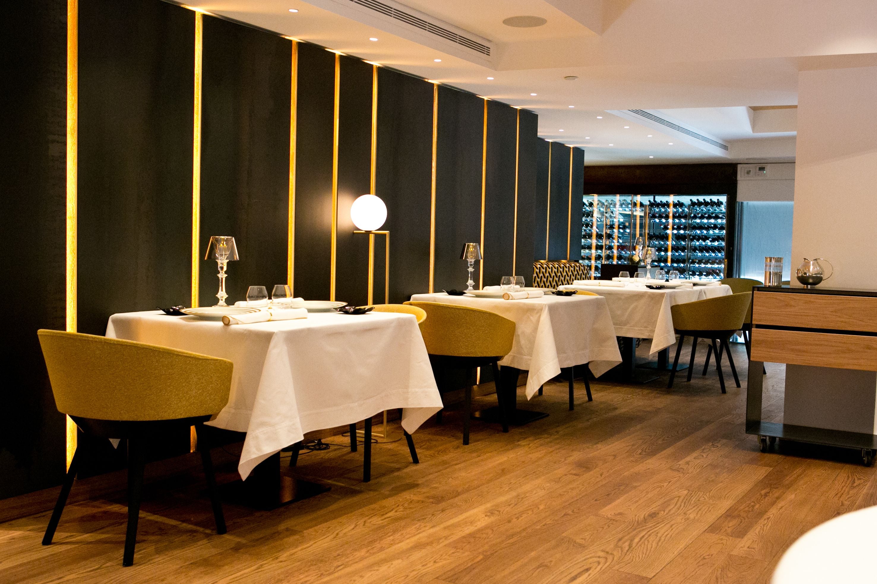 A restaurant with tables set for dining, featuring a modern interior with wooden flooring and dark walls accented by vertical light fixtures.