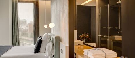 A hotel room with a bed, bedside table with a glass of water, a towel, a robe, a mirror, a shower area, and a view of the city through the window.