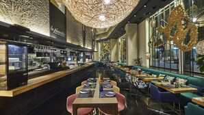 A modern restaurant interior with a long counter, multiple tables, and a high ceiling with decorative lighting.